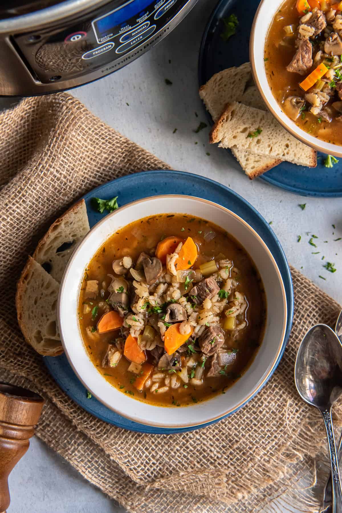 A top down shot of a bowl of beef barley soup with a slow cooker in the background.