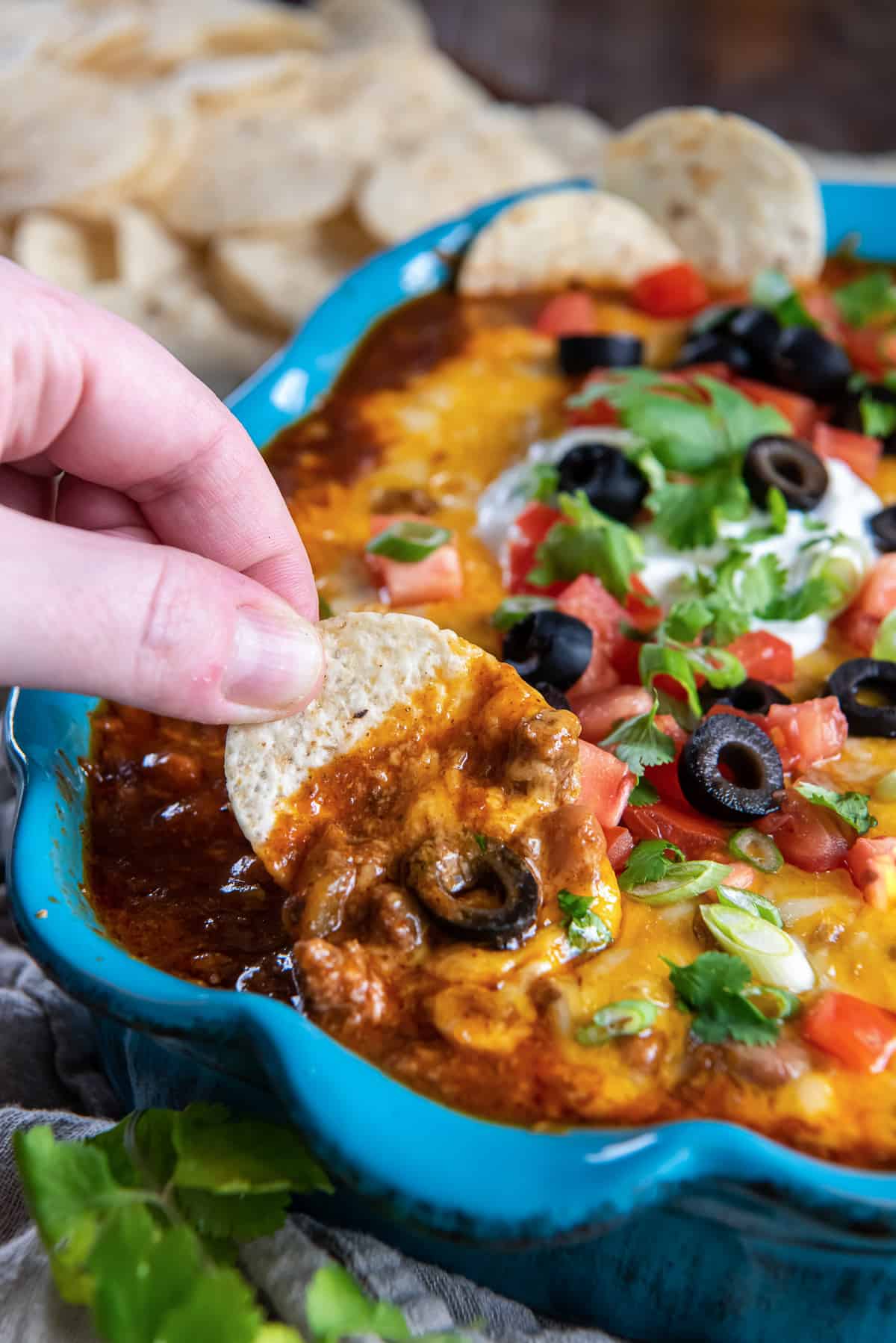 A hand dipping a tortilla chip into cheesy beef enchilada dip in a blue baking dish.