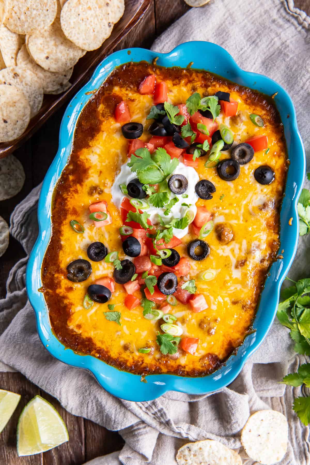 Beef enchilada dip in a blue baking dish topped with melted cheese, olives, and sour cream surrounded by lime wedges and tortilla chips.