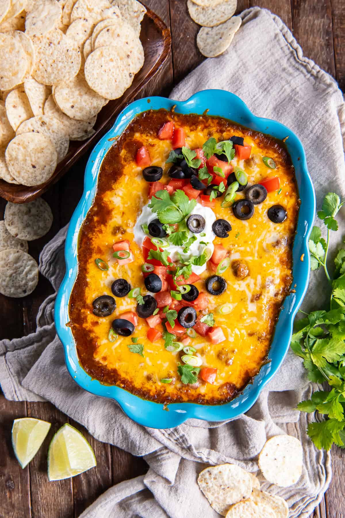 Beef enchilada dip in a blue baking dish topped with melted cheese, olives, and sour cream surrounded by lime wedges and tortilla chips.