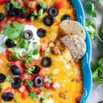 Two tortilla chips pressing into a blue baking dish filled with cheesy beef enchilada dip.