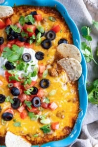 Two tortilla chips pressing into a blue baking dish filled with cheesy beef enchilada dip.