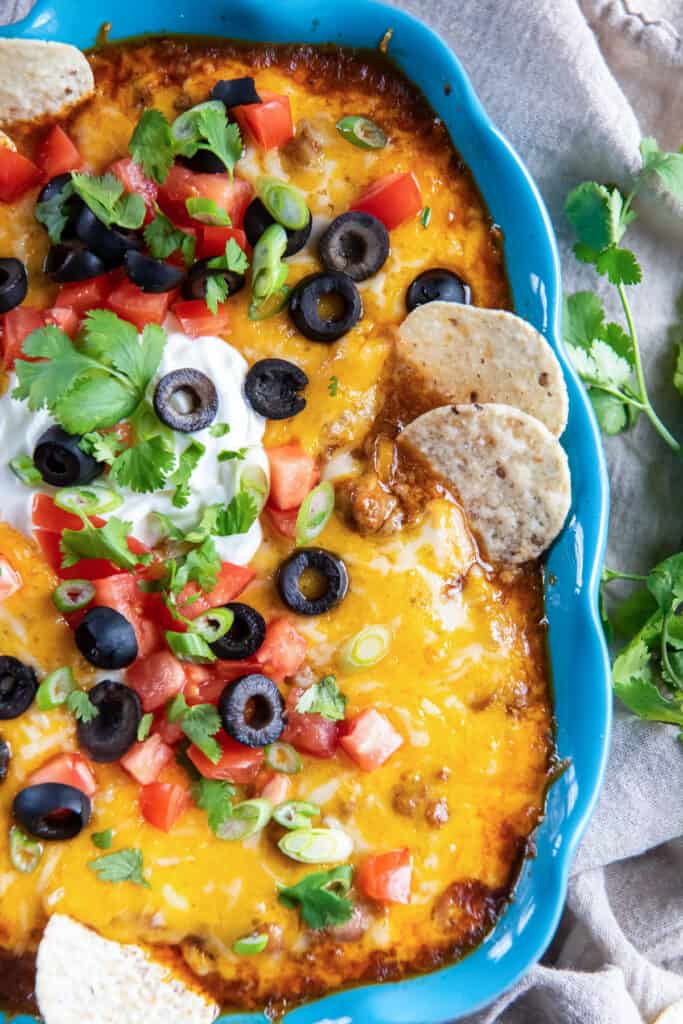 Two tortilla chips pressing into a blue baking dish filled with cheesy beef enchilada dip.