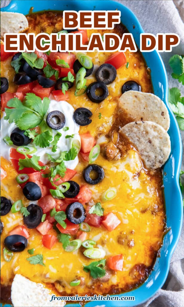 Two tortilla chips pressing into a blue baking dish filled with cheesy beef enchilada dip with text.