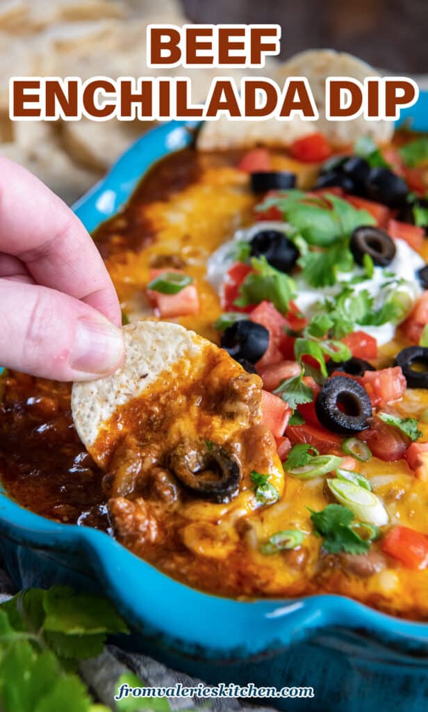 A hand dipping a tortilla chip into cheesy beef enchilada dip in a blue baking dish with text.