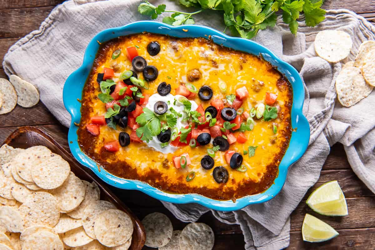 Beef enchilada dip in a blue baking dish topped with melted cheese, olives, and sour cream surrounded by lime wedges and tortilla chips.
