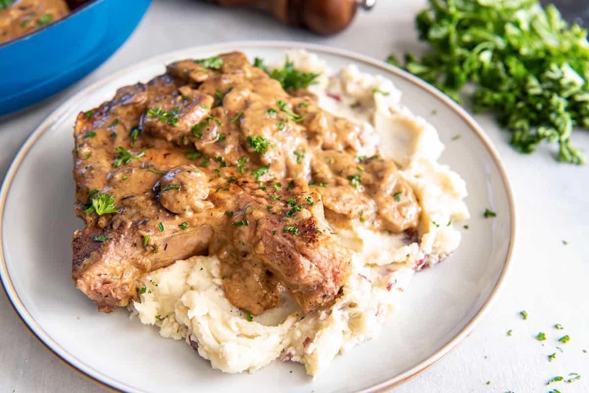 A mound of mashed potatoes topped with a pork chop with mushroom gravy on a dinner plate.