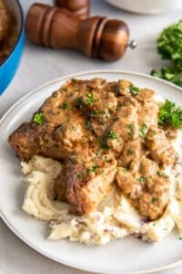 A pork chop topped with mushroom gravy over a mound of mashed potatoes on a dinner plate.