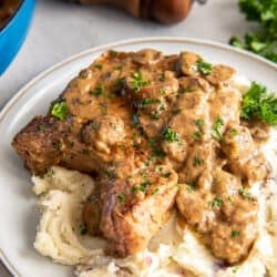 A pork chop topped with mushroom gravy over a mound of mashed potatoes on a dinner plate.