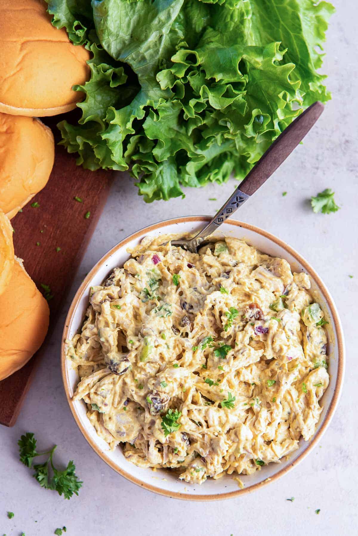 Curry chicken salad in a bowl with a spoon next to sandwich rolls and lettuce.