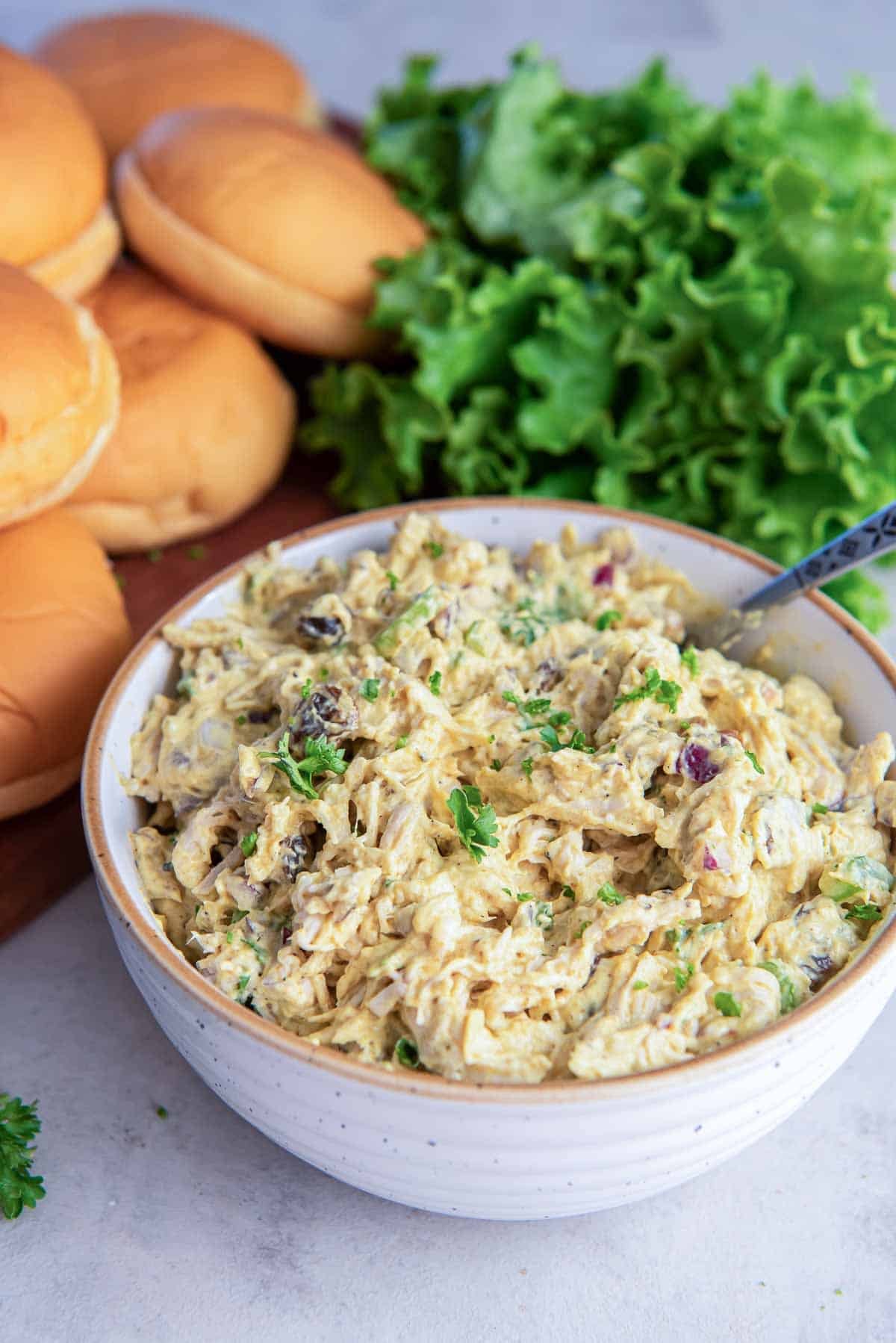 Curry chicken salad in a bowl with a spoon next to sandwich rolls and lettuce.