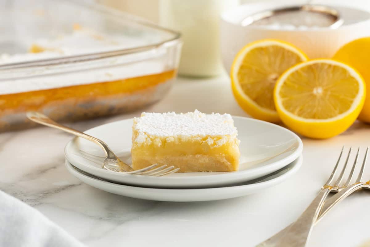A lemon bar on a stack of two white plates with cut lemons in the background.