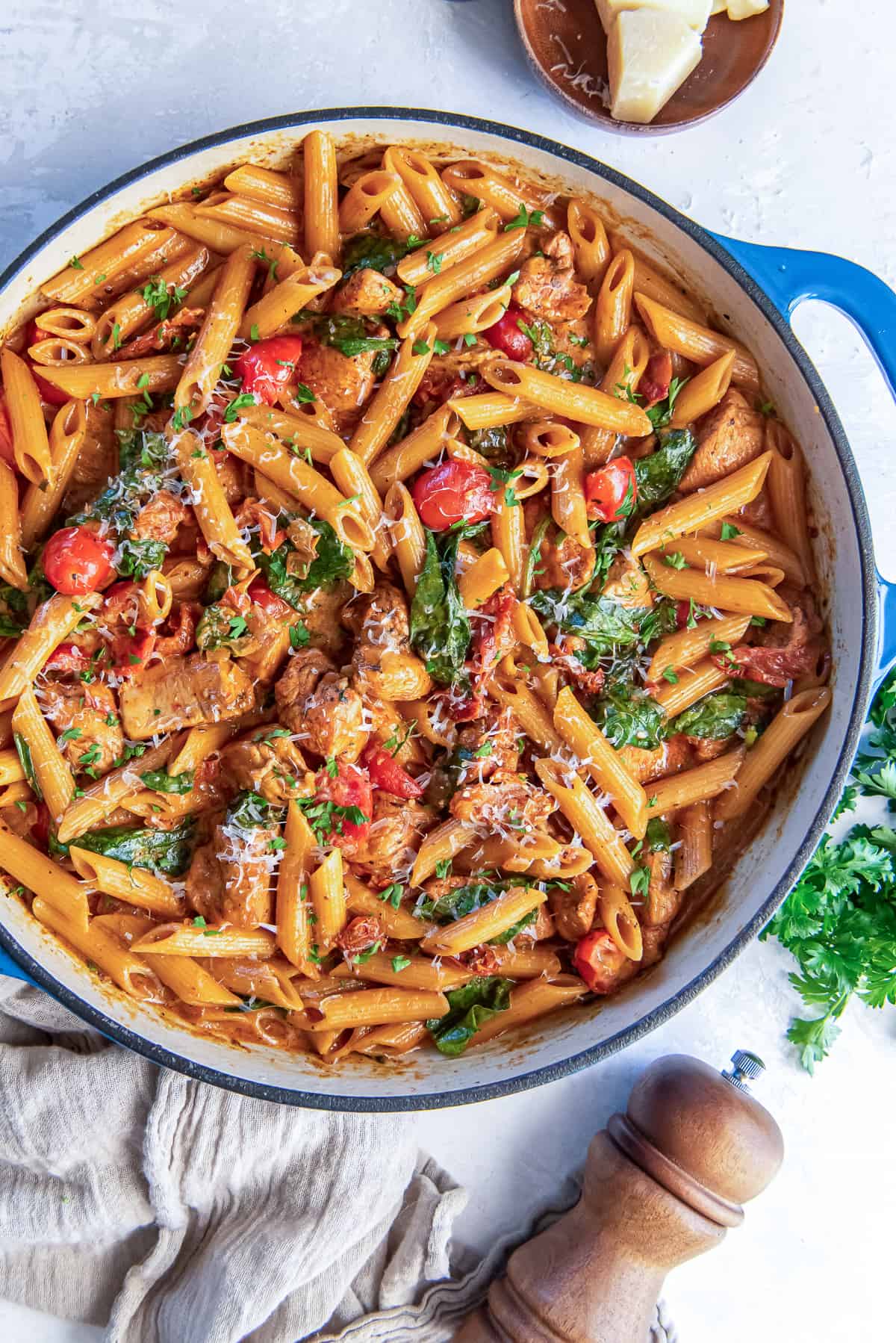 A skillet full of pasta with chicken, sundried tomatoes, cherry tomatoes, and spinach next to a pepper grinder.