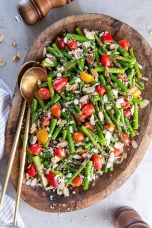 Asparagus salad with cherry tomatoes, almonds, and feta on a wood platter.