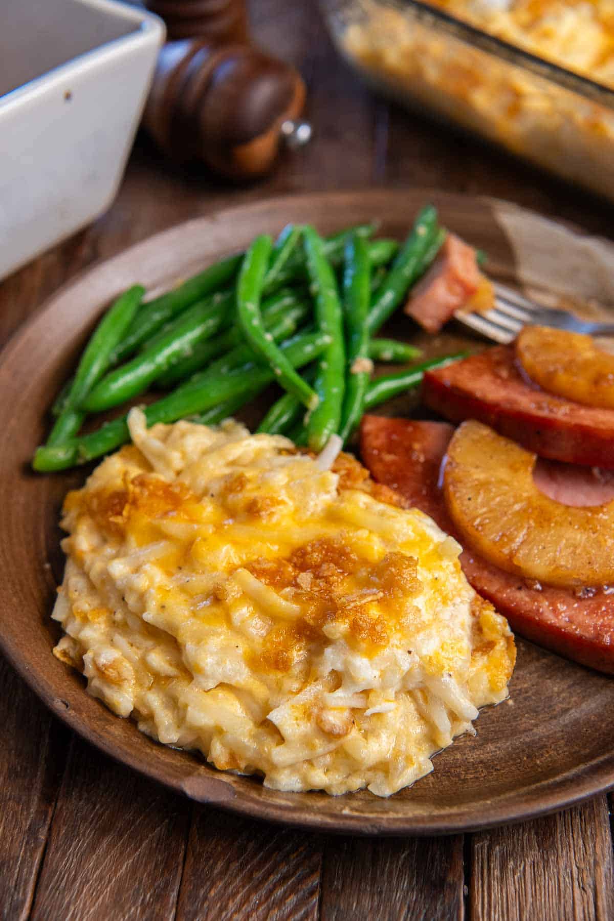 A serving of hash brown casserole on a brown plate with ham and green beans.