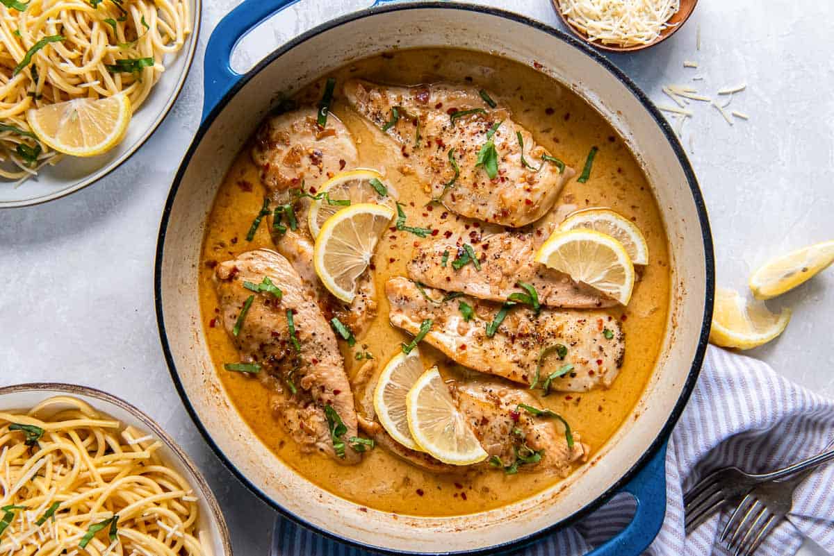 A large blue pan contains several pieces of cooked chicken breast in a creamy sauce, garnished with lemon slices and fresh basil. Surrounding the pan are plates of spaghetti, a bowl of grated cheese, and a striped napkin with utensils.