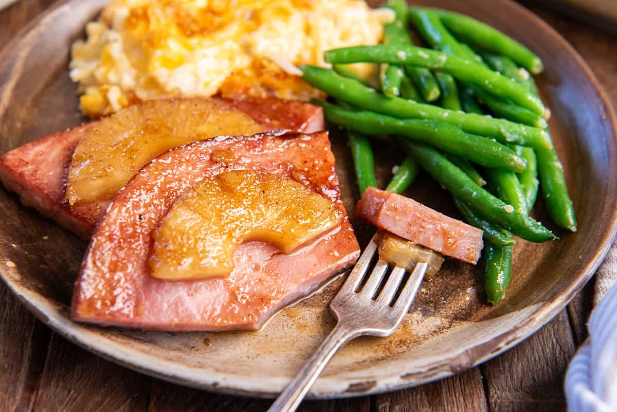A fork resting on a plate with pineapple ham steaks with cheesy hash browns and green beans.