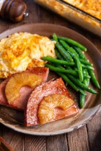 Pineapple glazed ham steak on a brown plate with cheesy hash browns and green beans.