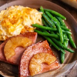 Pineapple glazed ham steak on a brown plate with cheesy hash browns and green beans.