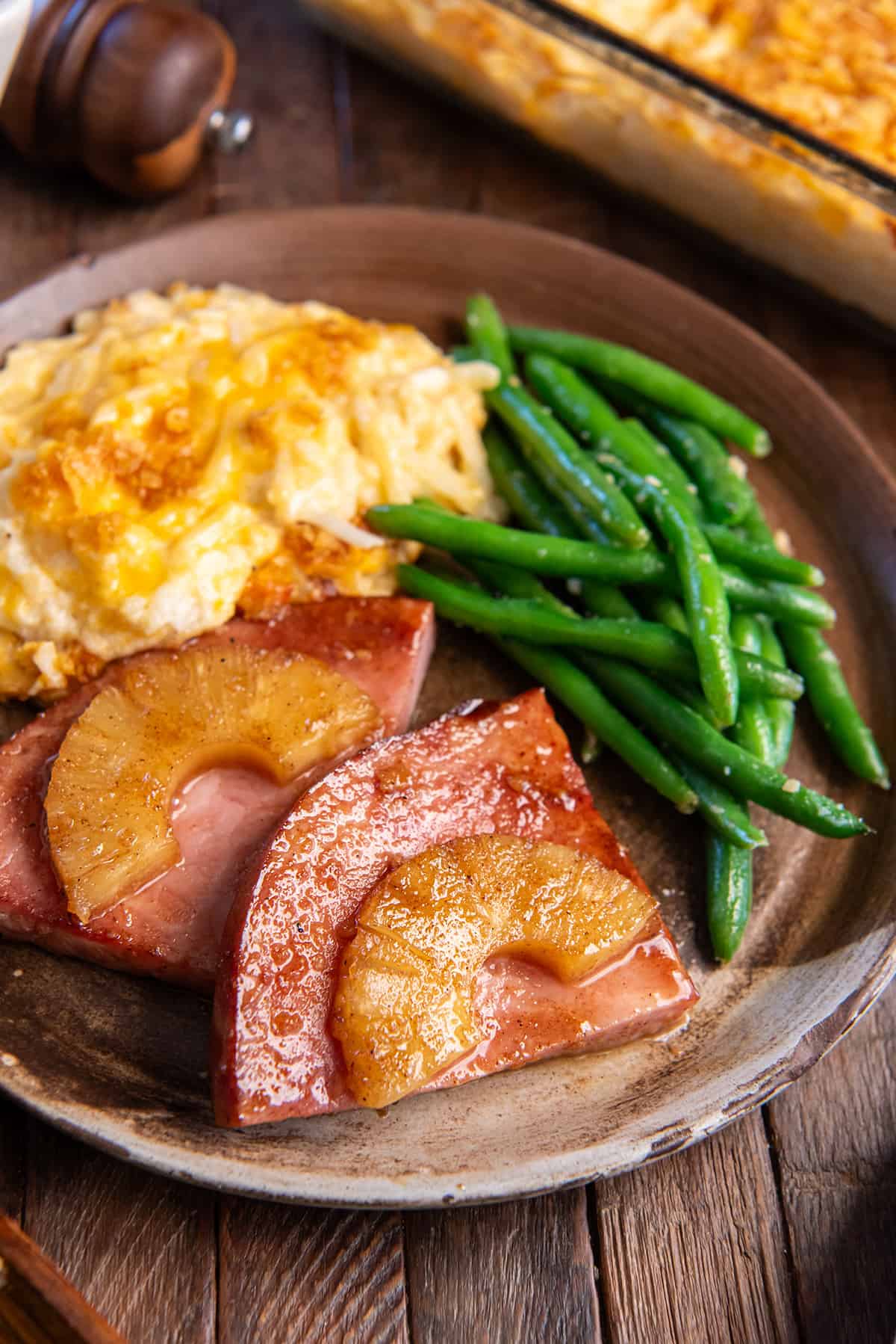 Pineapple glazed ham steak on a brown plate with cheesy hash browns and green beans.