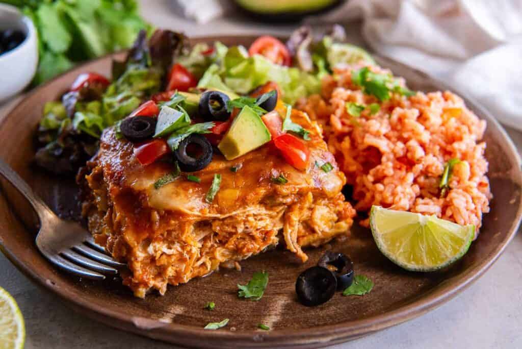 A wooden plate with a serving of cheesy casserole topped with diced tomatoes, avocado, and black olives. It's accompanied by a portion of rice, a fresh salad, and a lime wedge. A fork rests on the plate.