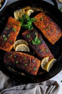 Four blackened salmon fillets in a cast iron skillet with lemon slices.