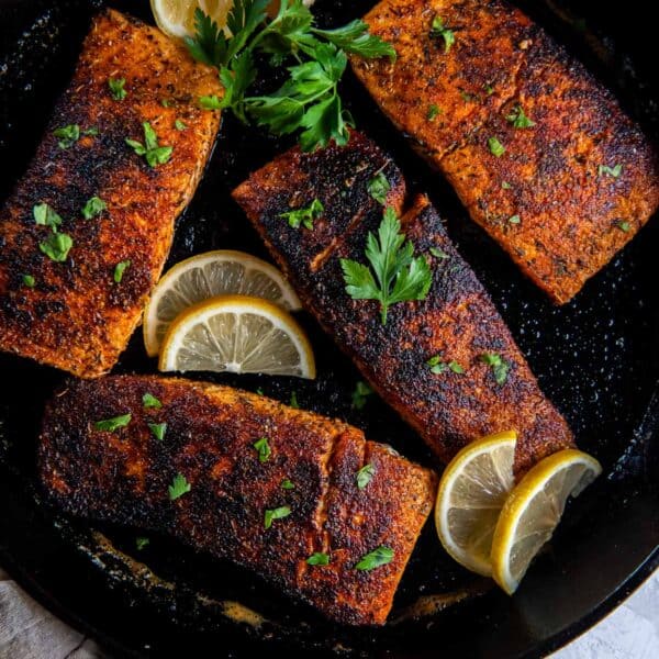 Four blackened salmon fillets in a cast iron skillet with lemon slices.