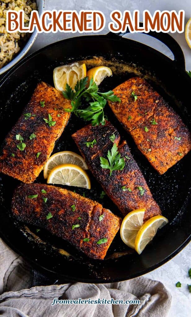 Four blackened salmon fillets in a cast iron skillet with lemon slices with text.