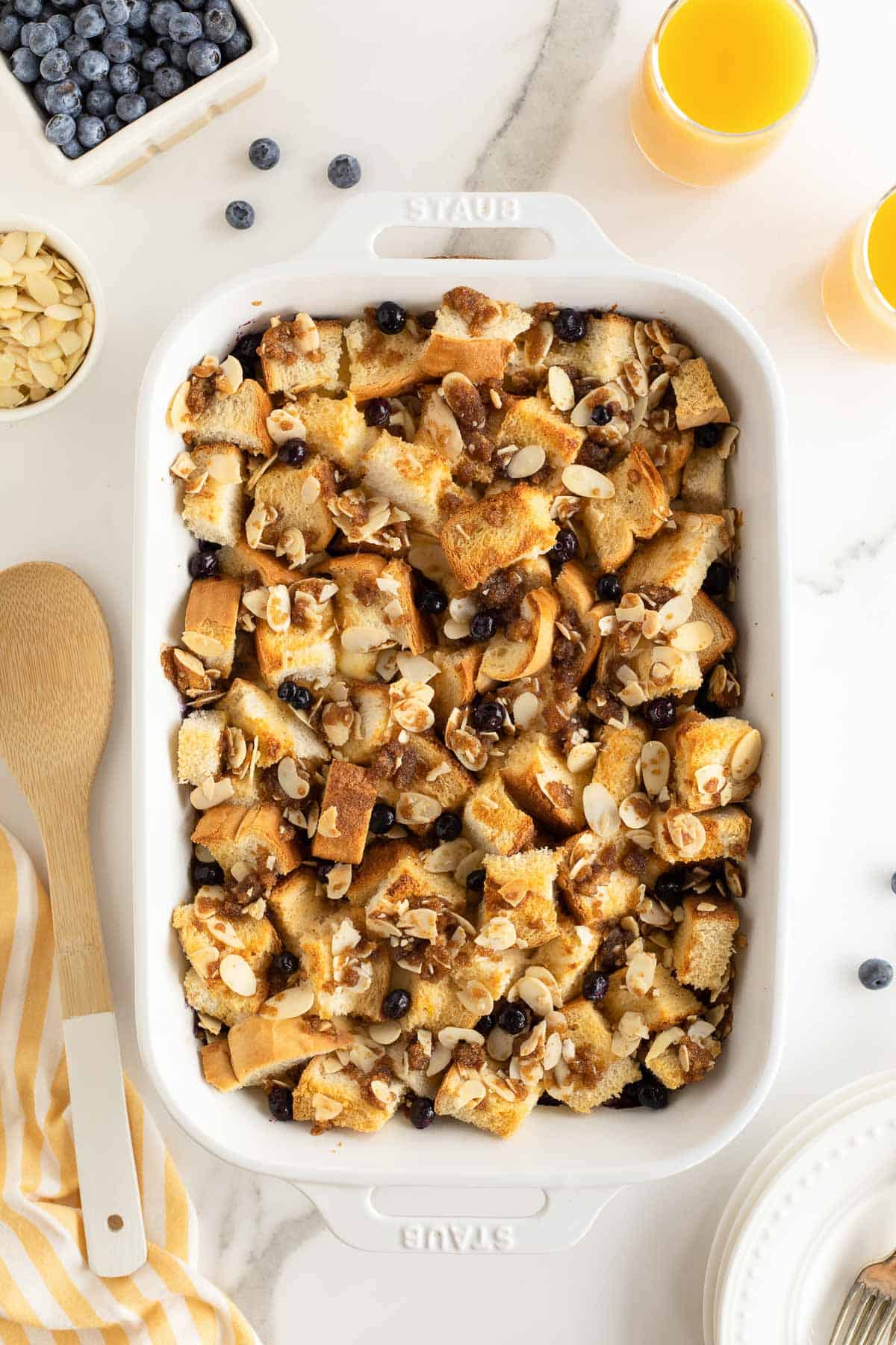 Blueberry French toast casserole in a white baking dish surrounded by glasses of orange juice and a carton of blueberries.