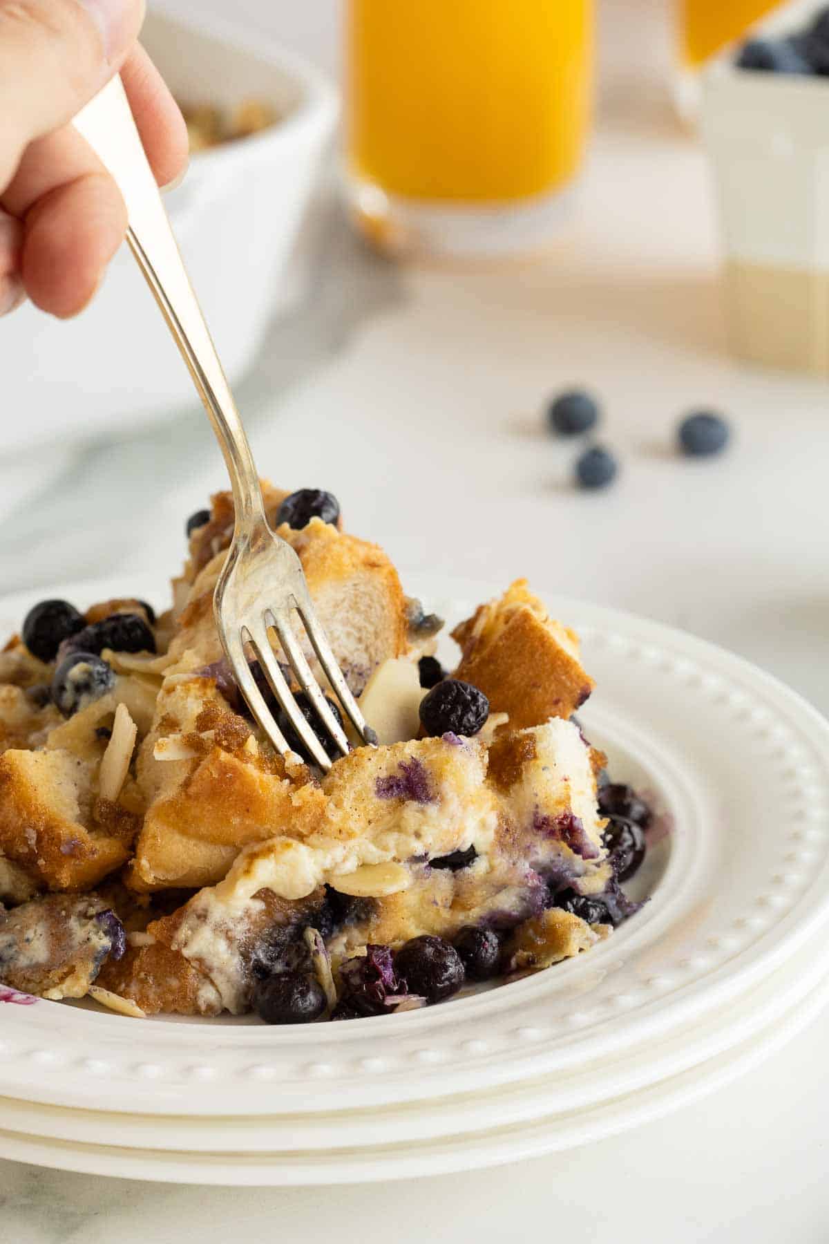 A fork pressing into a serving of blueberry French Toast casserole on a stack of white plates.