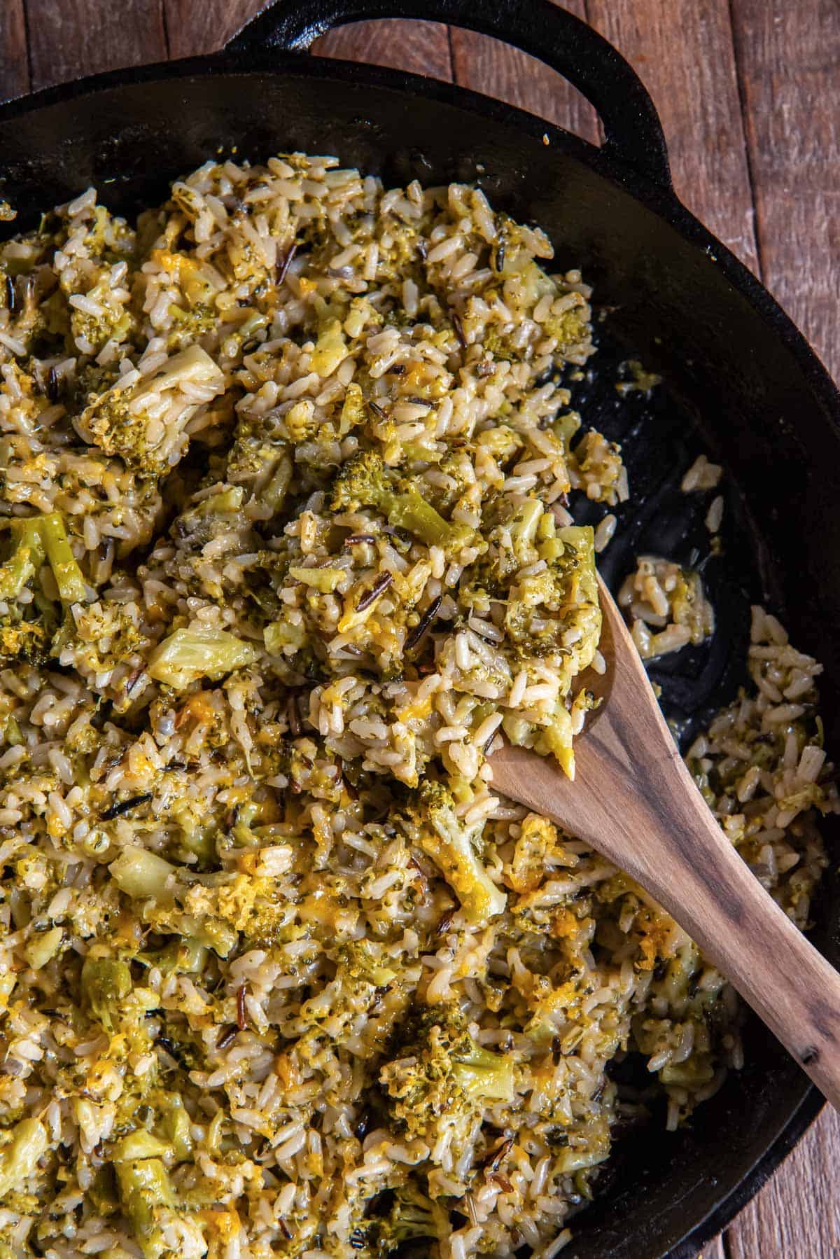 A wooden spoon scooping broccoli cheese rice from a cast iron skillet.