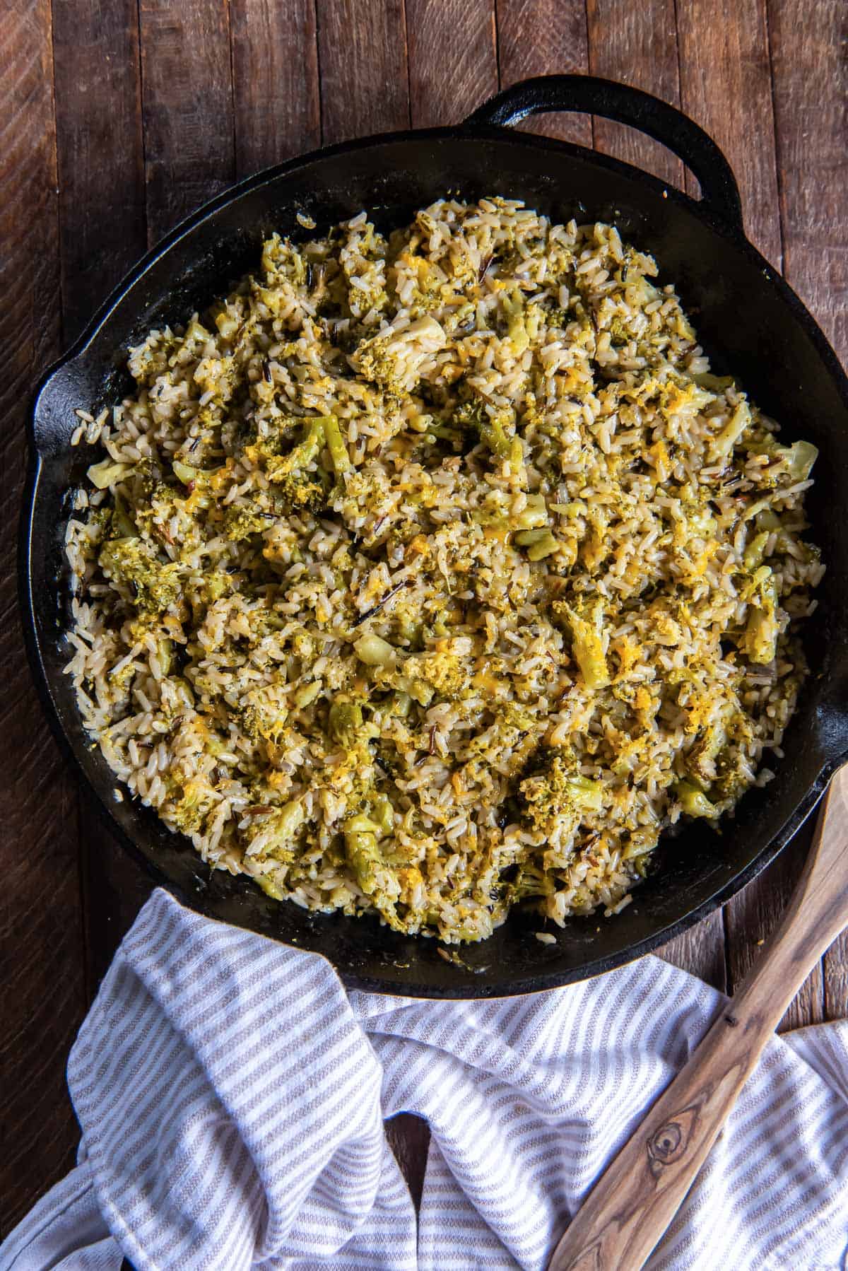 A wooden spoon and a striped kitchen cloth lying next to a skillet of broccoli cheese rice.