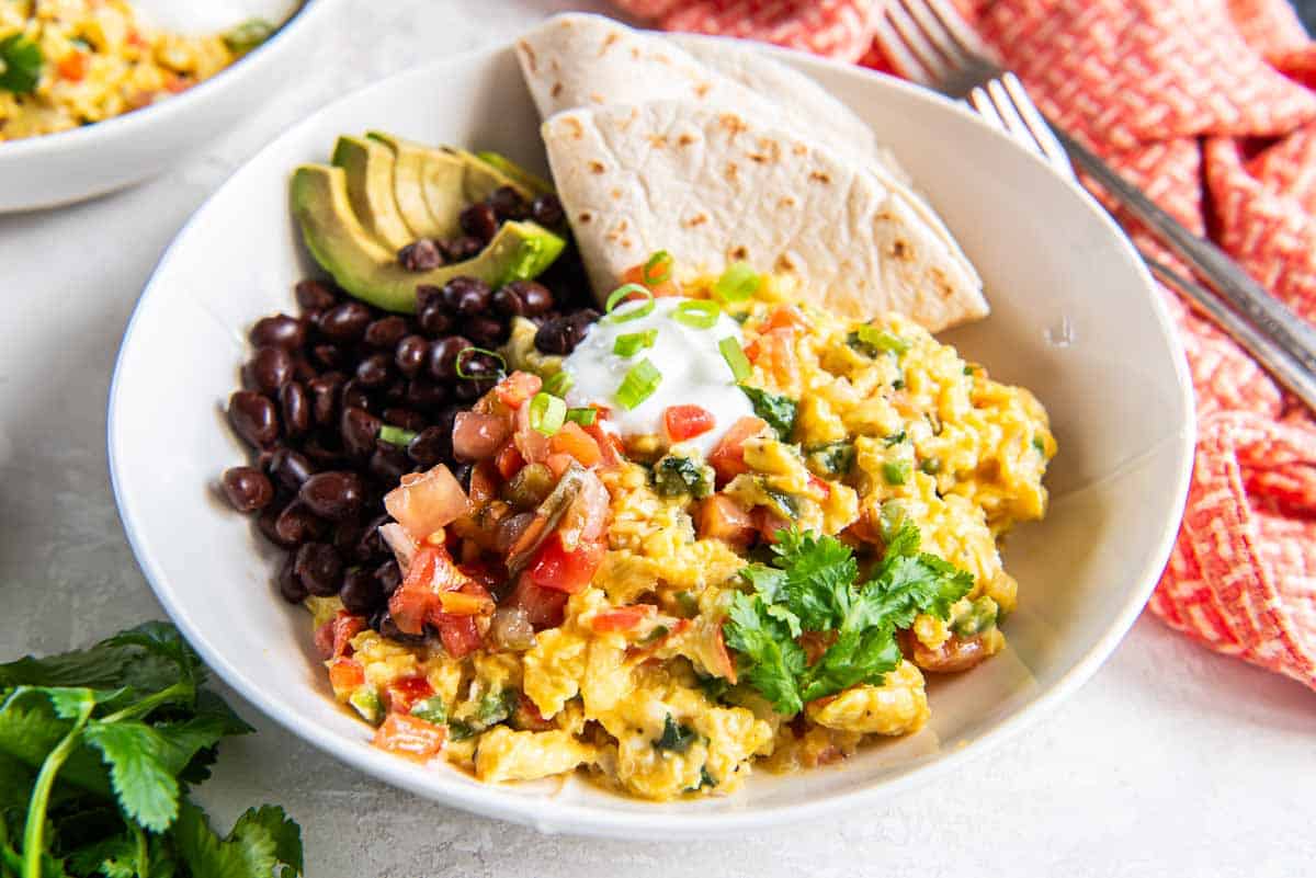 Mexican scrambled eggs in a white bowl with black beans, salsa, sour cream, and a folded flour tortilla.