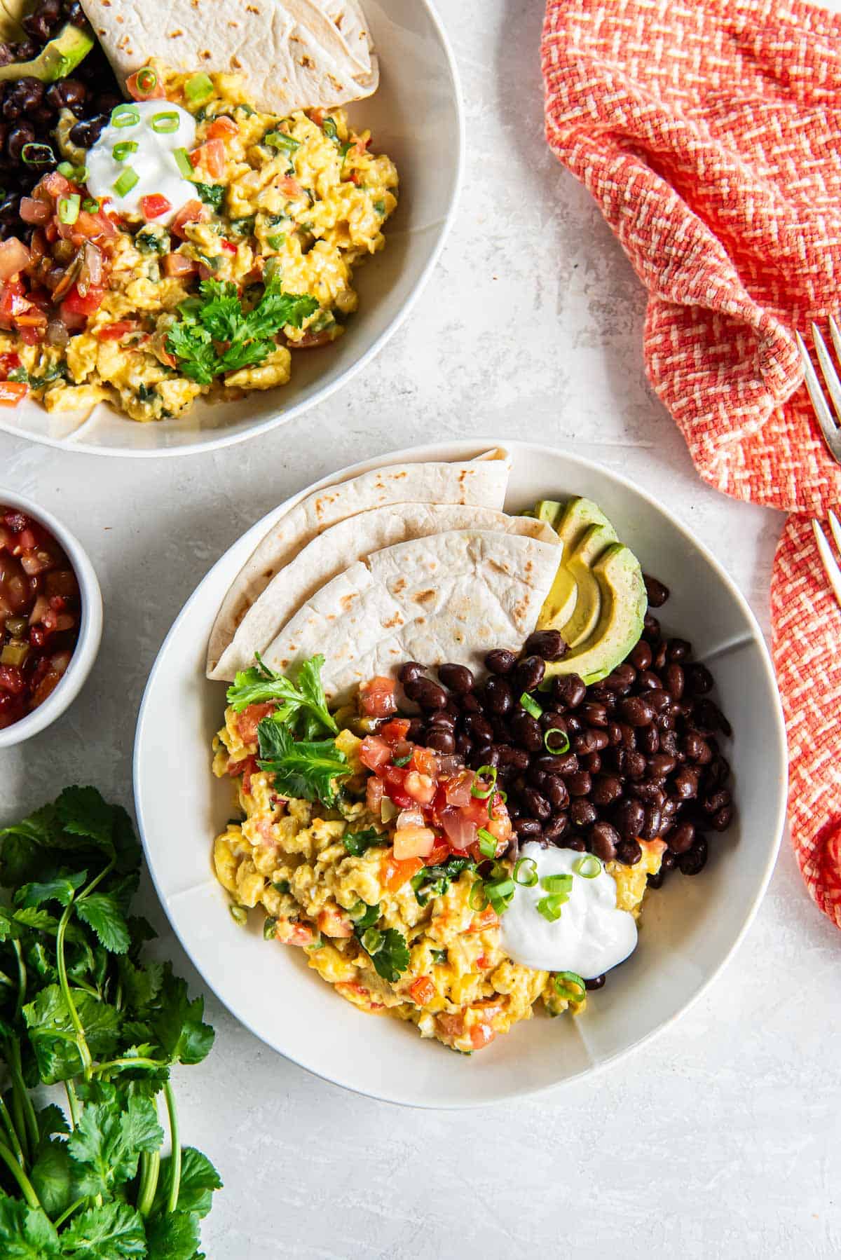 Two bowls filled with Mexican scrambled eggs, black beans, sliced avocado, and flour tortillas.