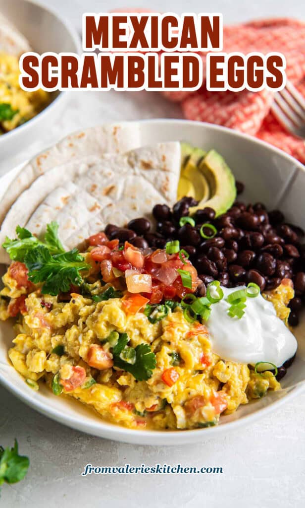 Mexican scrambled eggs in a white bowl with black beans, salsa, sour cream, and a folded flour tortilla with text.