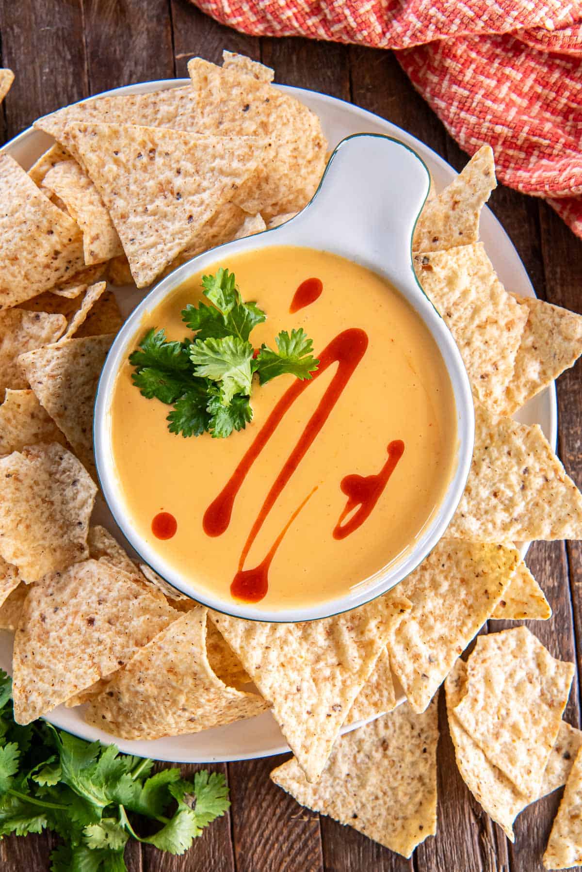 Nacho cheese sauce in a small white dish topped with hot sauce and cilantro and surrounded by tortilla chips.