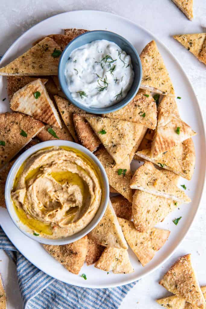 Homemde pita chips on a platter with small bowls of tzatziki sauce and hummus.