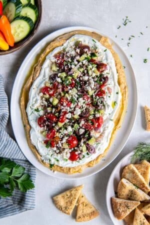 Layered Greek Dip on a white plate surrounded by vegetables and pita chips.
