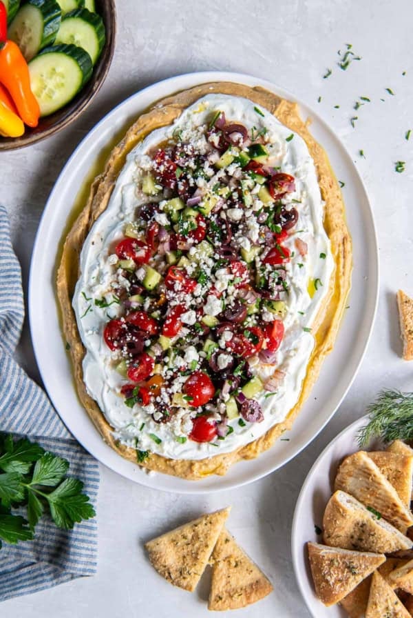 Layered Greek Dip on a white plate surrounded by vegetables and pita chips.