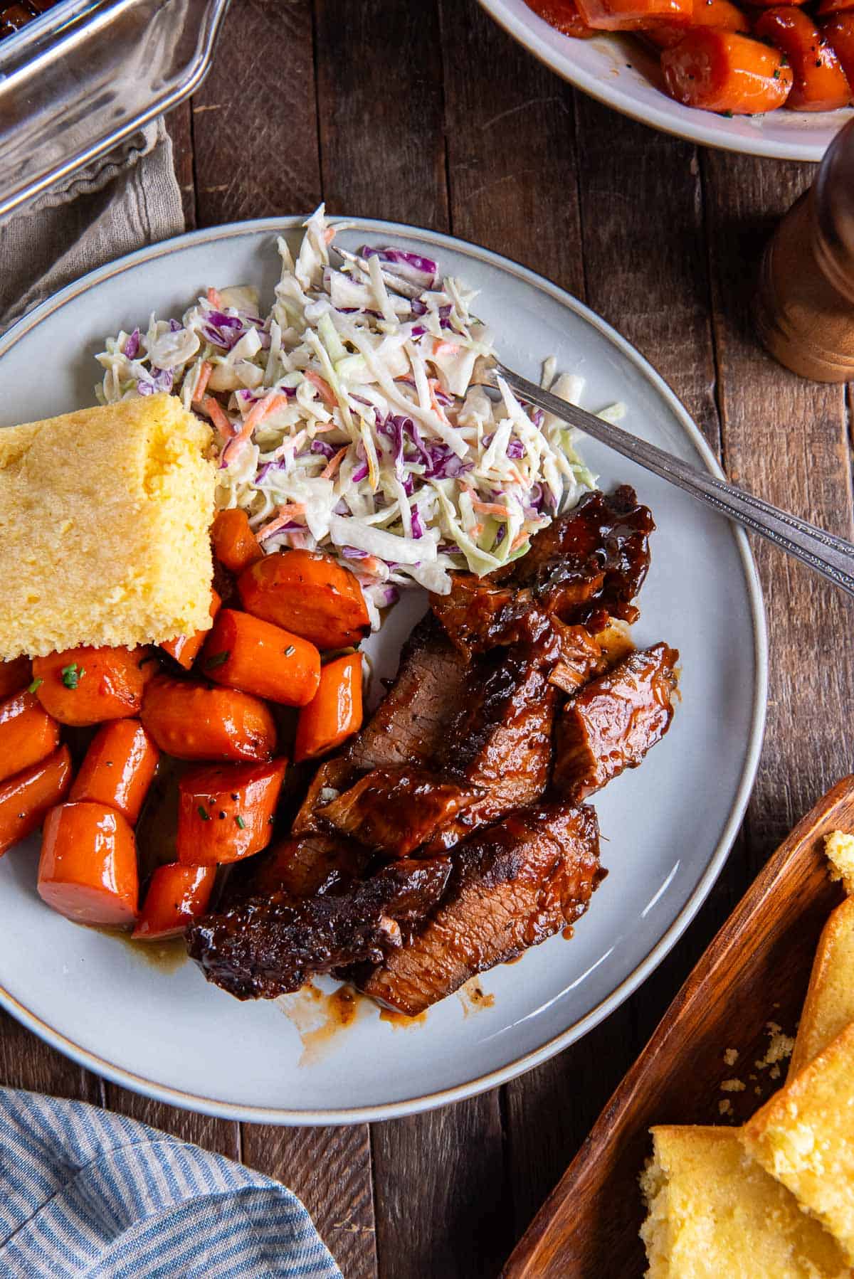 A serving of brisket on a dinner plate with carrots, coleslaw, and corned bread.