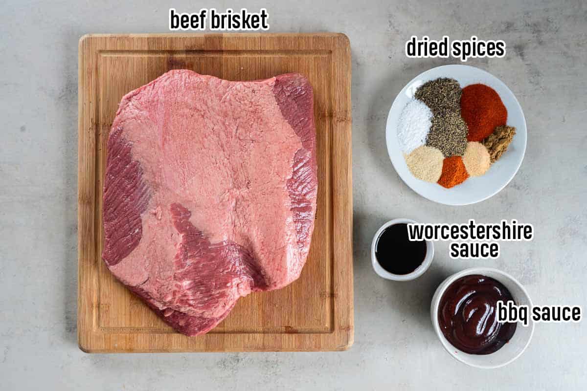 A beef brisket on a wood board next to a bowl of dried spices, worcestershire sauce, and bbq sauce.