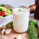 Buttermilk ranch dressing topped with fresh dill in a small mason jar on a cutting board.