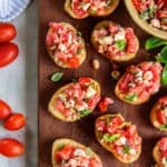 Sliced baguette topped with feta bruschetta on a brown board with a bowl full of bruschetta mixture.