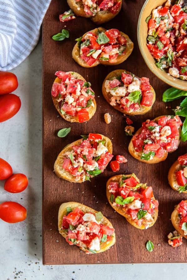 Sliced baguette topped with feta bruschetta on a brown board with a bowl full of bruschetta mixture.