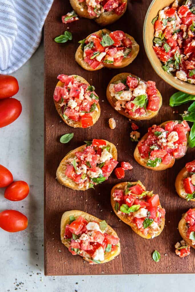 Sliced baguette topped with feta bruschetta on a brown board with a bowl full of bruschetta mixture.