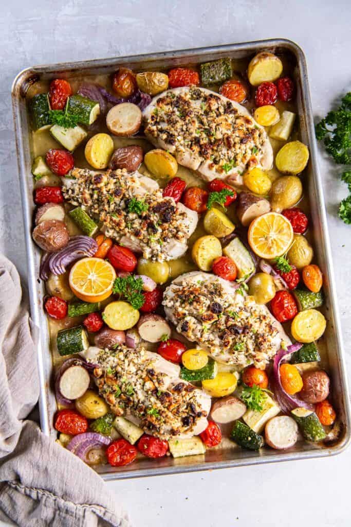 Greek feta stuffed chicken on a sheet pan with vegetables and lemon halves.