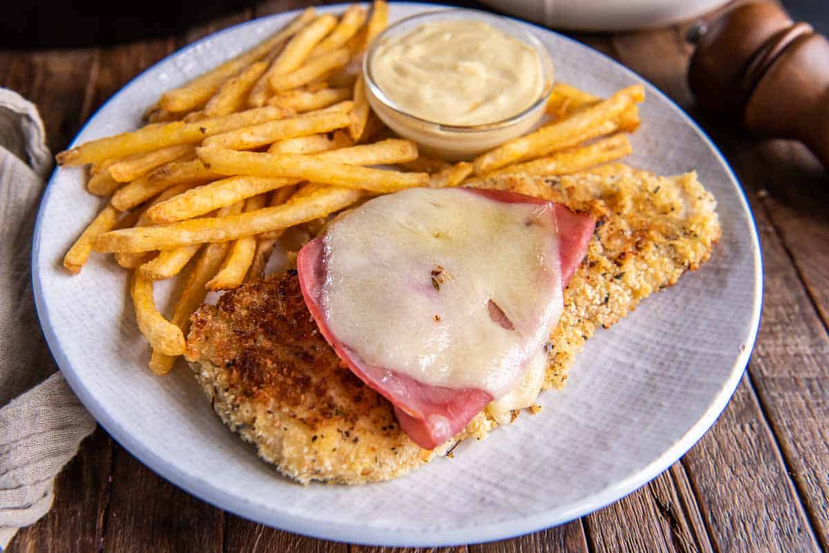 A plate with breaded and fried chicken breast topped with sliced ham and melted cheese, accompanied by crispy French fries. A small dish of creamy dipping sauce is also on the plate. The meal is served on a light-colored plate on a rustic wooden table.