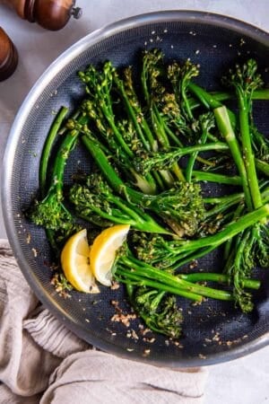 Sauteed broccolini in a skillet with lemon wedges.