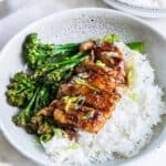 Sliced soy garlic chicken on top of white rice in a bowl with broccolini.