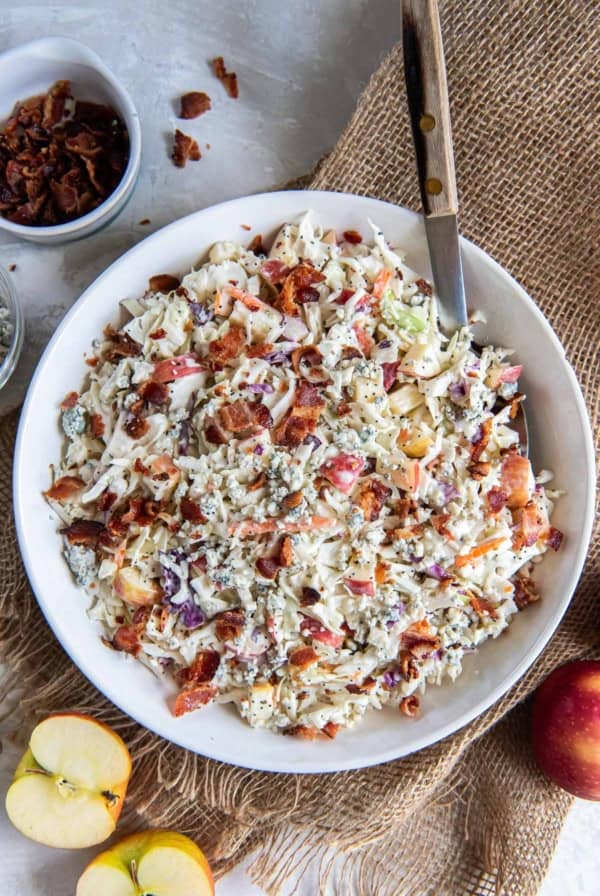 A bowl of apple coleslaw on a counter next to a small bowl of crumbled bacon and a halved apple.